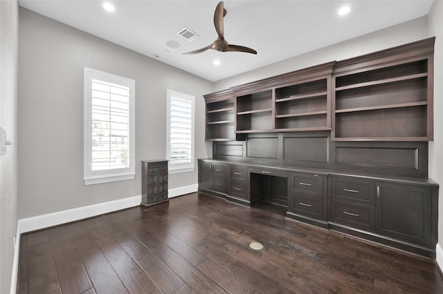 unfurnished office featuring dark wood-style floors, built in desk, visible vents, ceiling fan, and baseboards