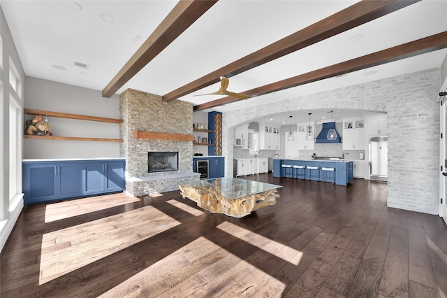 living area with arched walkways, dark wood-style flooring, visible vents, a stone fireplace, and beamed ceiling