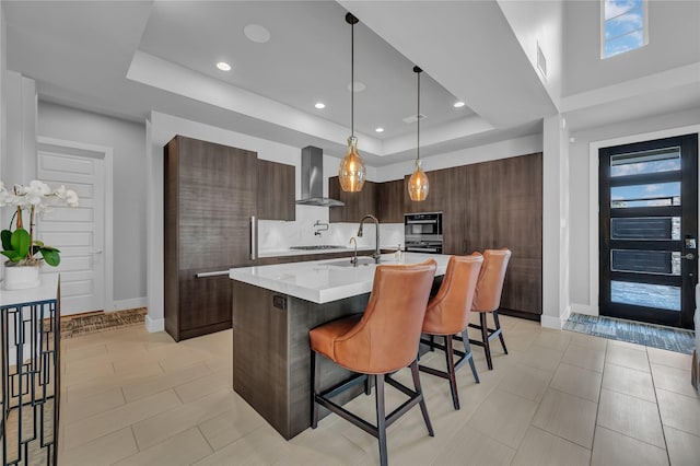 kitchen with wall chimney range hood, a breakfast bar area, dark brown cabinetry, a tray ceiling, and a center island with sink