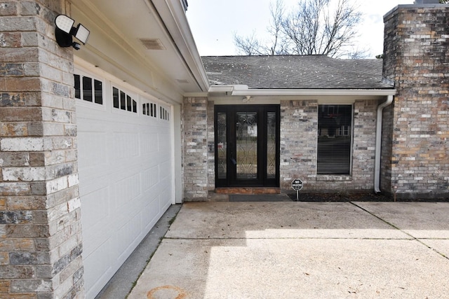 view of exterior entry with a garage, a shingled roof, brick siding, driveway, and a chimney