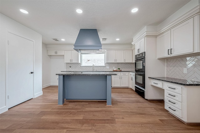 kitchen with white cabinetry, a center island, dark stone countertops, light wood-type flooring, and double oven