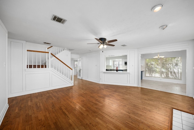 unfurnished living room featuring stairway, wood finished floors, visible vents, and ceiling fan with notable chandelier