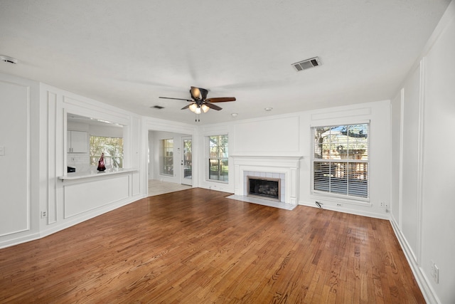unfurnished living room featuring a decorative wall, wood finished floors, ceiling fan, a tile fireplace, and visible vents