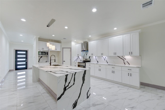 kitchen featuring wall chimney range hood, appliances with stainless steel finishes, an island with sink, white cabinets, and decorative light fixtures