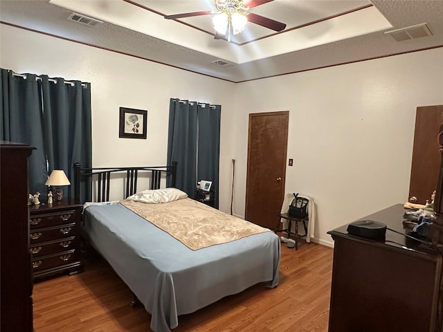 bedroom featuring a tray ceiling, wood finished floors, visible vents, and a ceiling fan