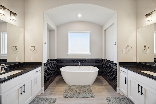 bathroom with a wainscoted wall, tile walls, two vanities, a sink, and a freestanding tub