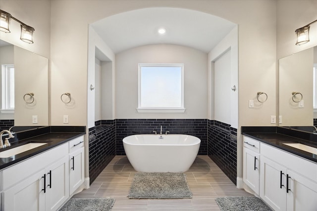 full bath with two vanities, a sink, tile walls, wainscoting, and a soaking tub