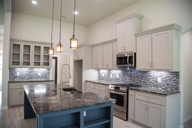 kitchen with a center island with sink, open shelves, appliances with stainless steel finishes, a sink, and dark stone countertops