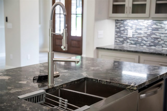 kitchen with a sink, white cabinetry, dark stone counters, tasteful backsplash, and glass insert cabinets