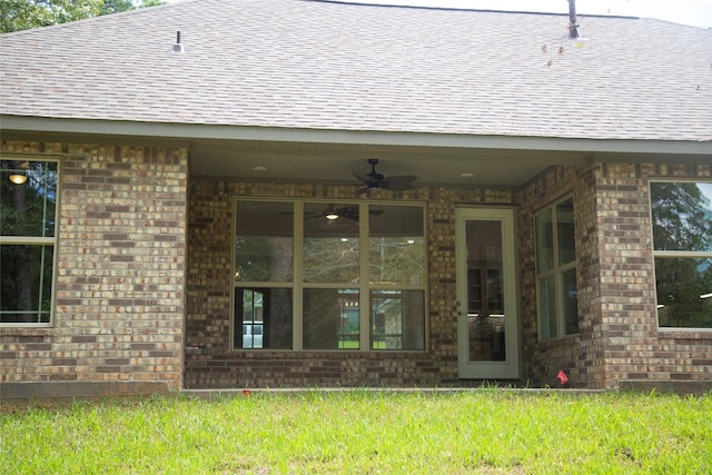 entrance to property featuring brick siding, a lawn, a shingled roof, and a ceiling fan