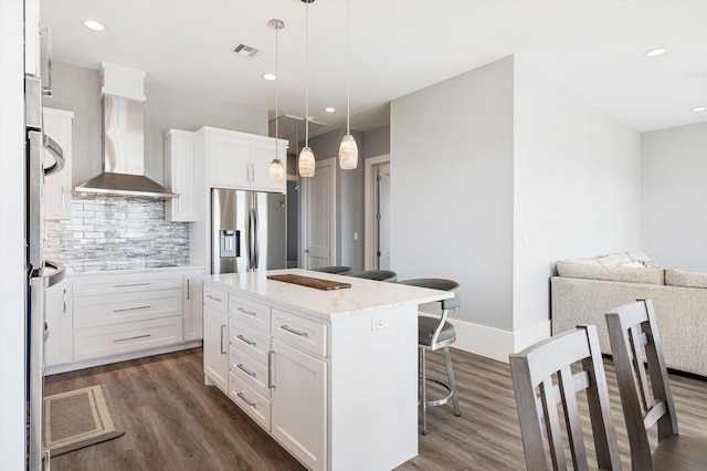 kitchen featuring wall chimney range hood, stainless steel fridge, a center island, white cabinets, and decorative light fixtures