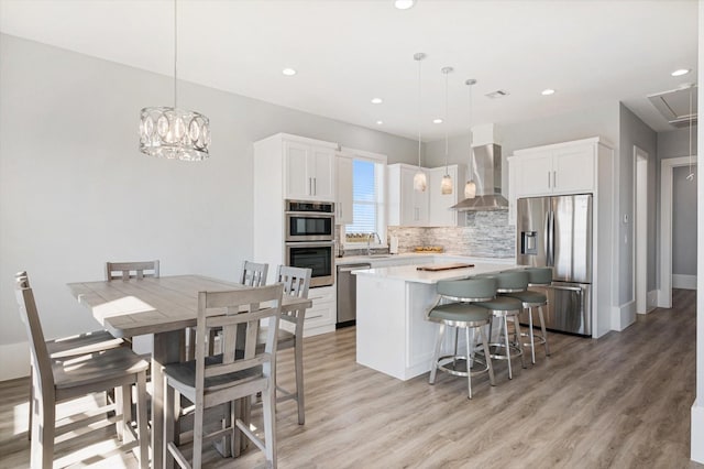 kitchen with appliances with stainless steel finishes, white cabinets, hanging light fixtures, a center island, and wall chimney range hood