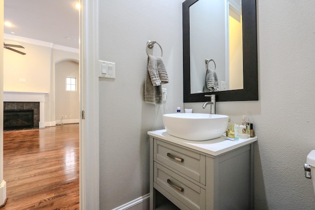 bathroom with a tiled fireplace, vanity, ornamental molding, hardwood / wood-style flooring, and ceiling fan