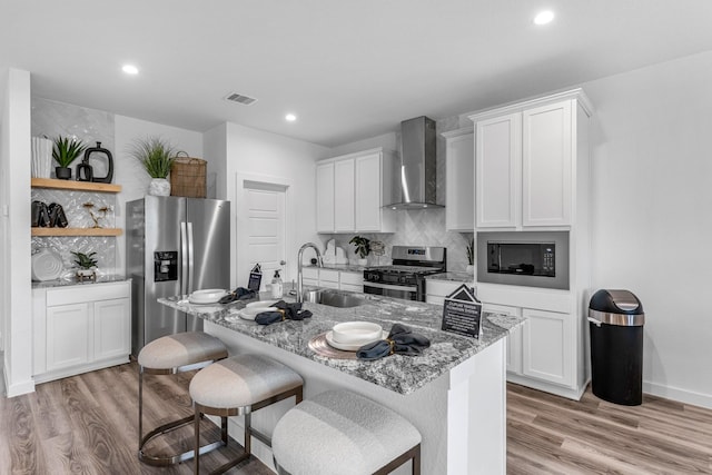 kitchen featuring white cabinetry, an island with sink, appliances with stainless steel finishes, and wall chimney range hood