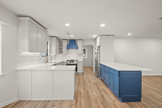 kitchen with wall chimney exhaust hood, blue cabinets, light wood-type flooring, appliances with stainless steel finishes, and kitchen peninsula