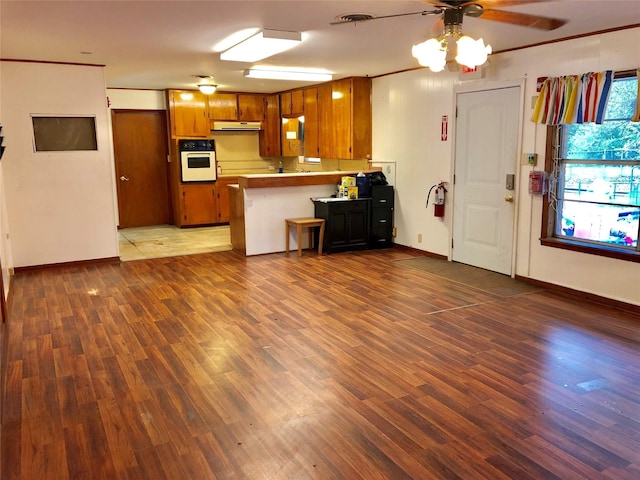 kitchen with dark wood-type flooring, ornamental molding, kitchen peninsula, ceiling fan, and oven