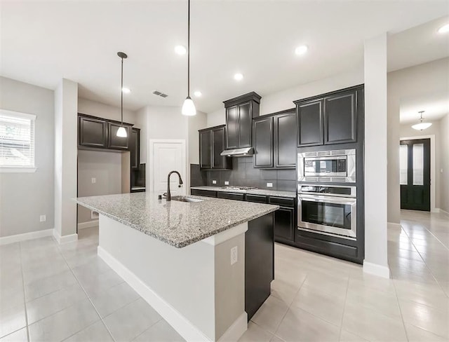 kitchen with sink, light stone counters, hanging light fixtures, appliances with stainless steel finishes, and a kitchen island with sink