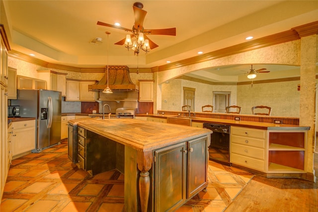 kitchen with butcher block countertops, ceiling fan, black appliances, custom range hood, and an island with sink