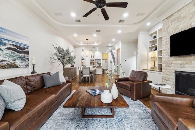 living room featuring dark hardwood / wood-style floors, a tray ceiling, ornamental molding, built in shelves, and a stone fireplace