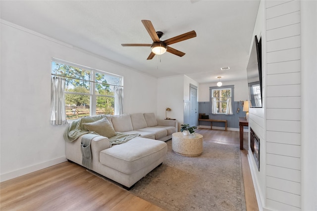 living area featuring wood finished floors, a ceiling fan, baseboards, ornamental molding, and a glass covered fireplace