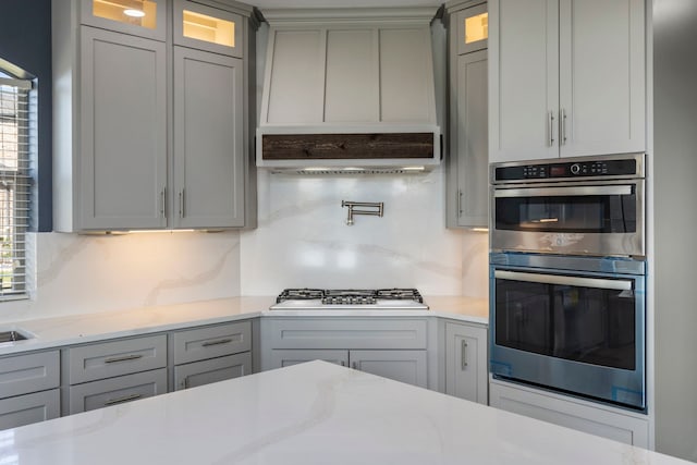 kitchen with appliances with stainless steel finishes, gray cabinetry, backsplash, light stone countertops, and custom range hood