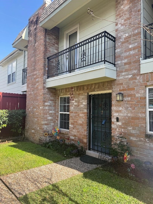 property entrance featuring a balcony, fence, a lawn, and brick siding