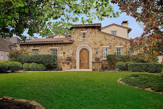 mediterranean / spanish-style home with a tile roof, stone siding, a front yard, and a chimney