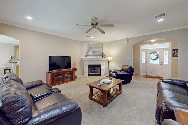 living room with ceiling fan, ornamental molding, a stone fireplace, and light carpet