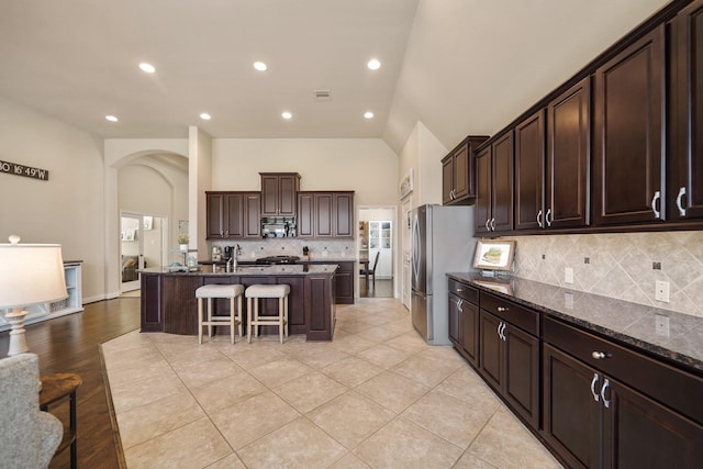 kitchen with stainless steel fridge, dark stone countertops, dark brown cabinetry, an island with sink, and a kitchen bar