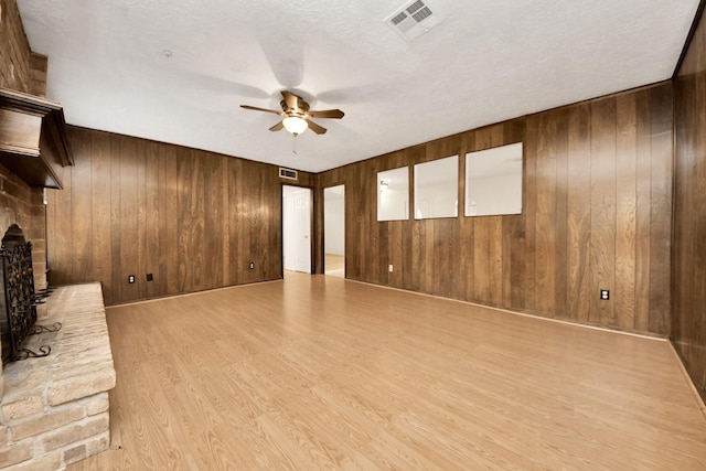 unfurnished living room featuring wooden walls, ceiling fan, a brick fireplace, a textured ceiling, and light hardwood / wood-style flooring