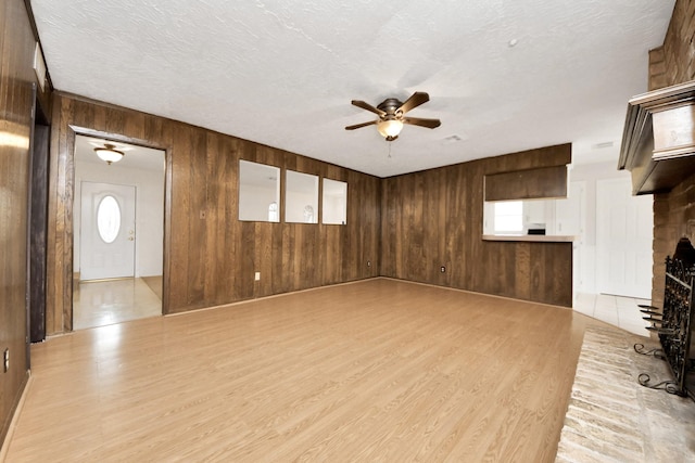 unfurnished living room with ceiling fan, a textured ceiling, a brick fireplace, light wood-type flooring, and wood walls