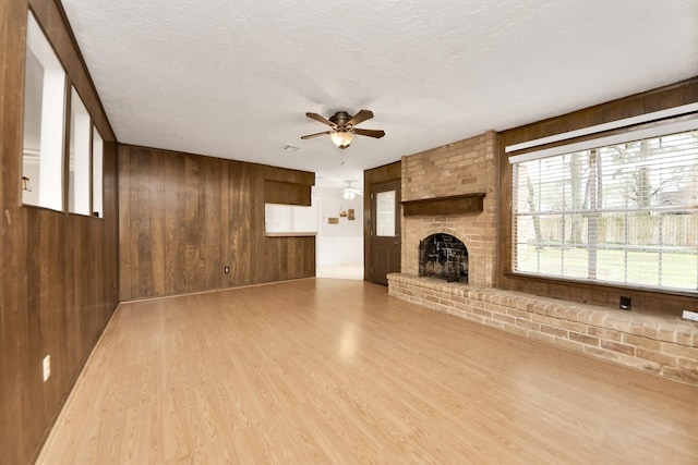 unfurnished living room featuring wood walls, wood-type flooring, a textured ceiling, a brick fireplace, and ceiling fan