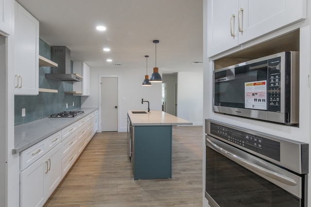 kitchen featuring stainless steel appliances, wall chimney range hood, a center island with sink, and white cabinets