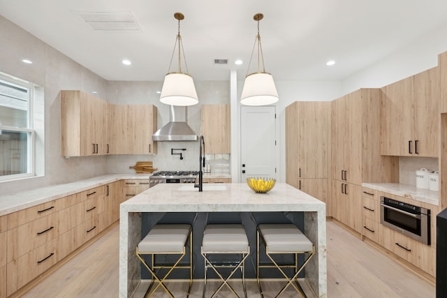 kitchen featuring stove, a center island with sink, oven, wall chimney exhaust hood, and light brown cabinets