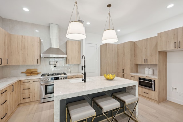 kitchen featuring wall chimney range hood, sink, a kitchen island with sink, stainless steel appliances, and light brown cabinets