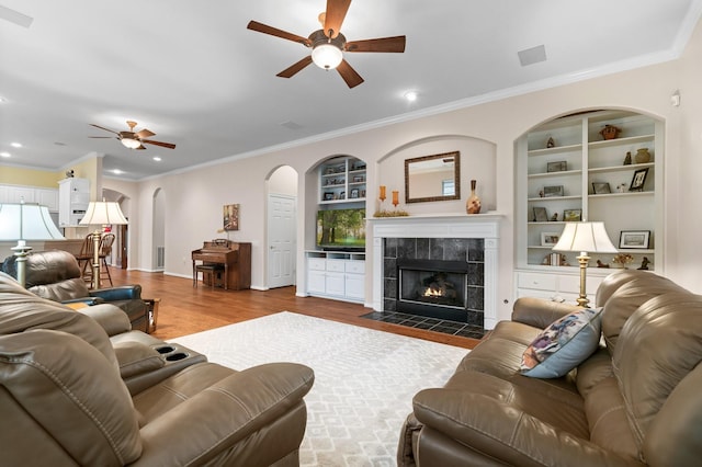 living room featuring built in shelves, crown molding, a tile fireplace, hardwood / wood-style flooring, and ceiling fan