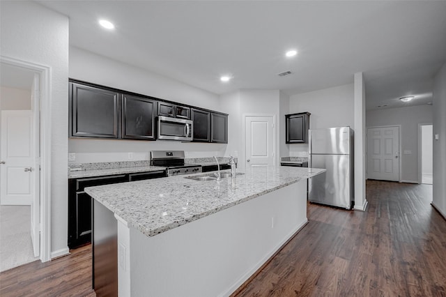 kitchen featuring sink, dark wood-type flooring, appliances with stainless steel finishes, light stone counters, and a center island with sink