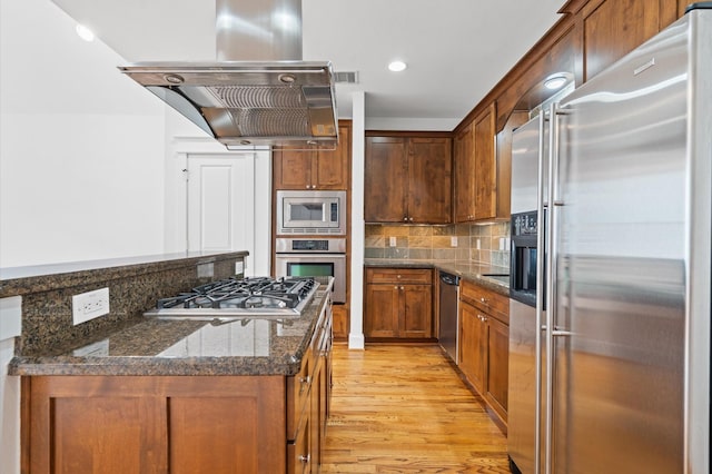 kitchen featuring stainless steel appliances, tasteful backsplash, light hardwood / wood-style floors, island exhaust hood, and dark stone counters