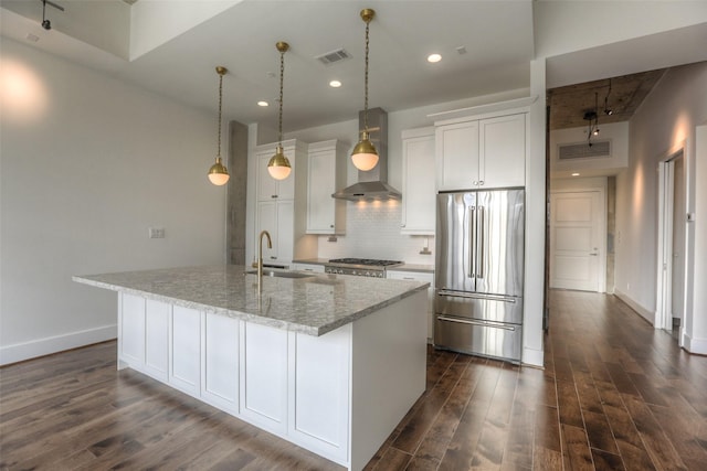 kitchen with sink, wall chimney range hood, stainless steel refrigerator, white cabinets, and a center island with sink