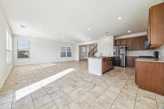 kitchen with an island with sink, stainless steel fridge, stove, light tile patterned floors, and light stone countertops