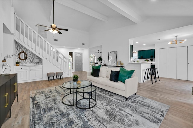 living room featuring light wood-type flooring, beamed ceiling, stairway, and ceiling fan with notable chandelier