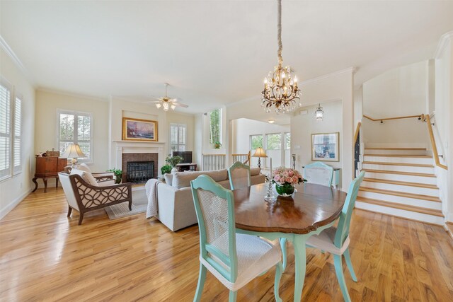 dining area featuring ceiling fan with notable chandelier, ornamental molding, a tiled fireplace, and light hardwood / wood-style floors