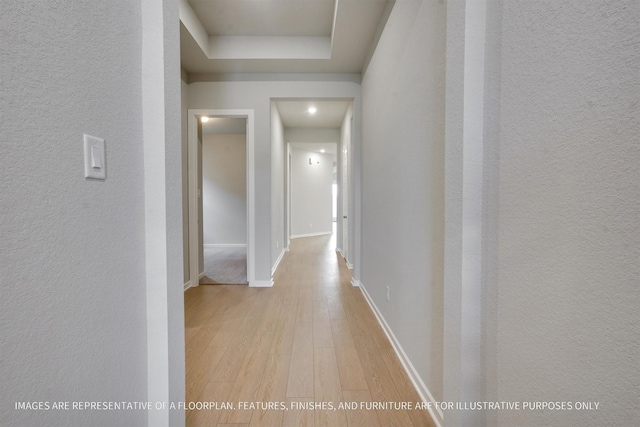 hallway with a textured wall, a tray ceiling, baseboards, and wood finished floors