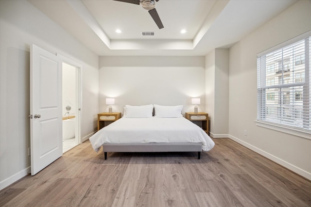 bedroom featuring ceiling fan, ensuite bathroom, a tray ceiling, and light hardwood / wood-style floors