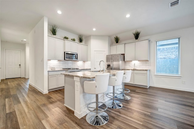 kitchen featuring appliances with stainless steel finishes, white cabinetry, a kitchen bar, light stone counters, and a center island with sink