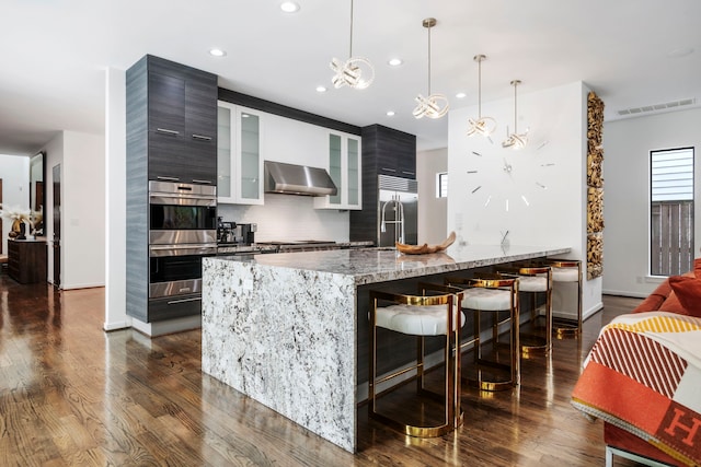 kitchen featuring a breakfast bar, appliances with stainless steel finishes, hanging light fixtures, light stone countertops, and wall chimney exhaust hood