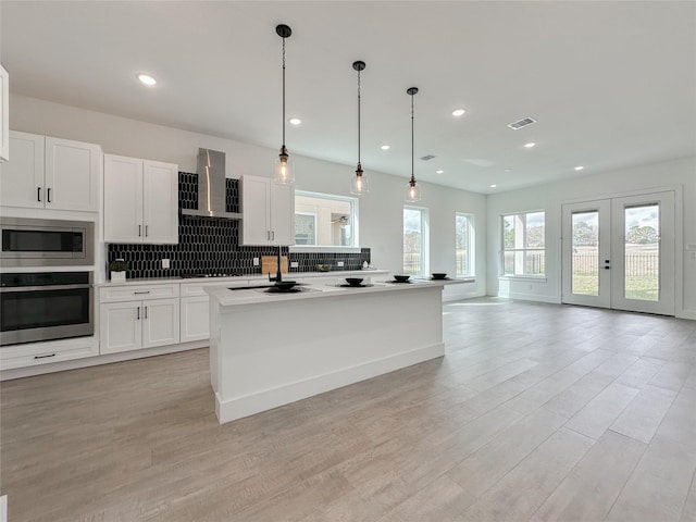 kitchen featuring wall chimney exhaust hood, appliances with stainless steel finishes, an island with sink, pendant lighting, and white cabinets