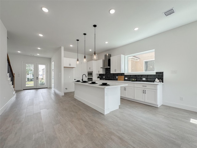 kitchen with wall chimney range hood, decorative light fixtures, white cabinets, and a center island with sink