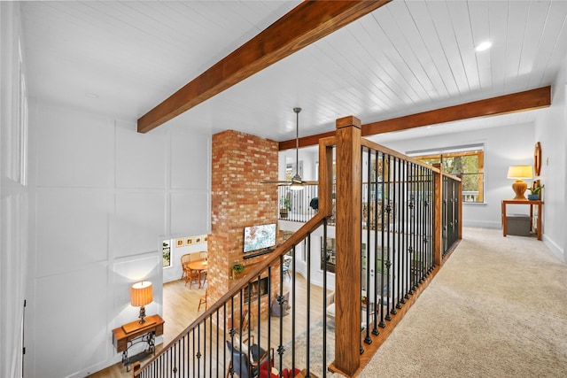 hallway featuring carpet, a decorative wall, an upstairs landing, beamed ceiling, and baseboards