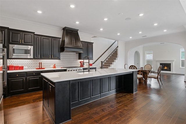 kitchen featuring a kitchen island with sink, crown molding, custom range hood, a sink, and stainless steel appliances